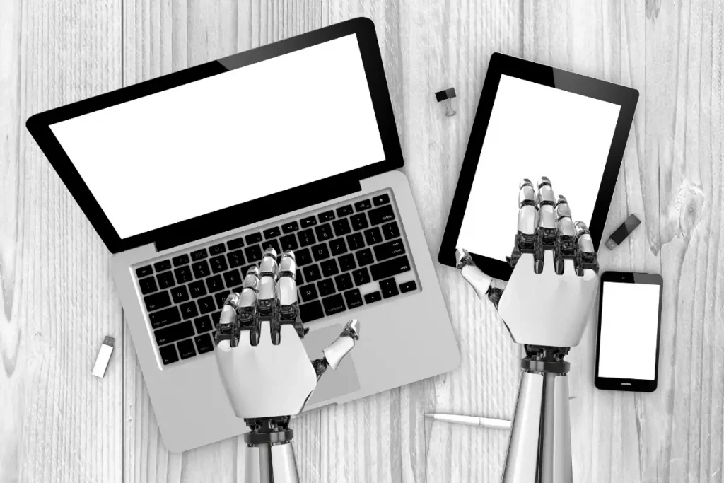 Duas mãos robóticas brancas e pretas digitando em um laptop e tablet sobre uma mesa de madeira representando o conceito de IA Agêntica